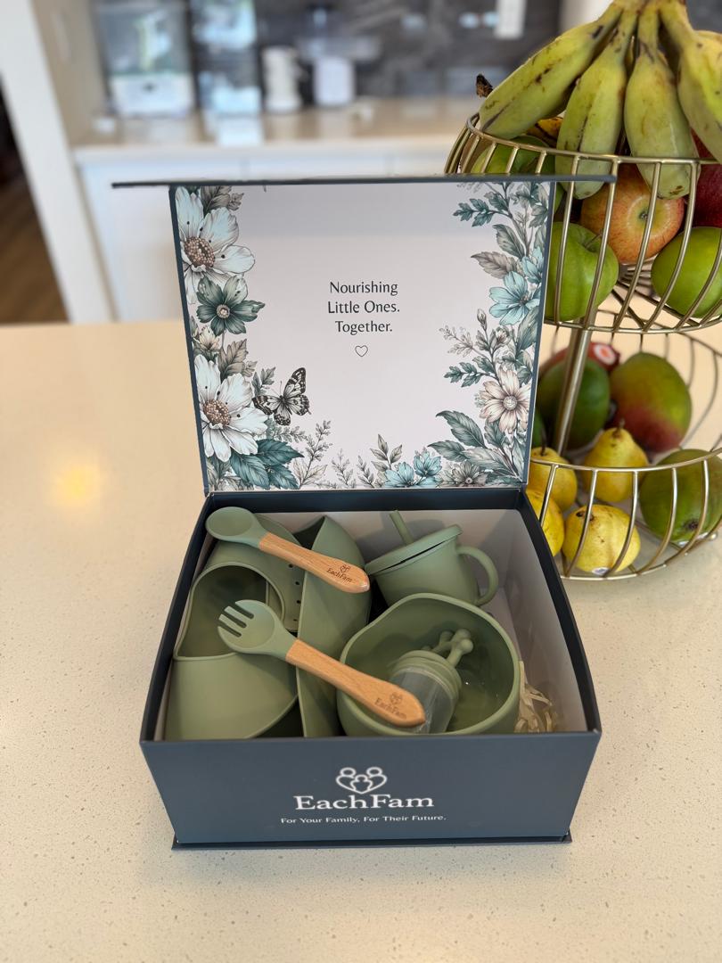 EachFam silicone feeding set gift box opened on a kitchen counter, showing sage-green bowl, cup, spoon and fork with wooden handles